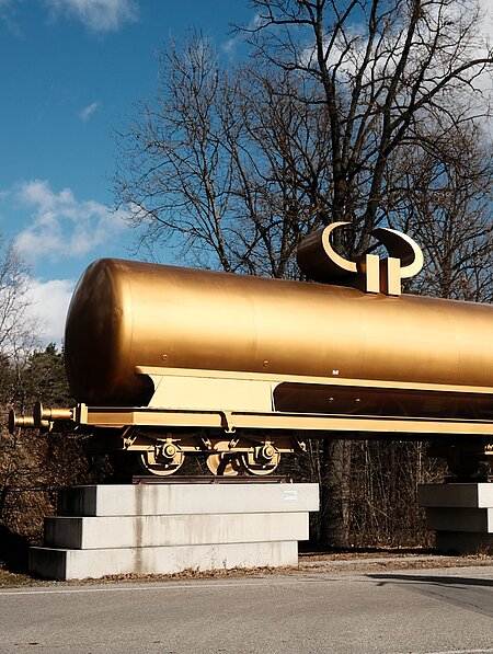 A golden tank wagon for transporting liquids such as mineral oil is decorated with a horizontal euro currency sign, which transforms the figure into a horned idol. It is located in front of the parking lot outside the park grounds and is the first sculpture that invites you to visit. 