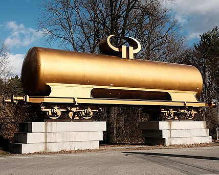 A golden tank wagon for transporting liquids such as mineral oil is decorated with a horizontal euro currency sign, which transforms the figure into a horned idol. It is located in front of the parking lot outside the park grounds and is the first sculpture that invites you to visit. 