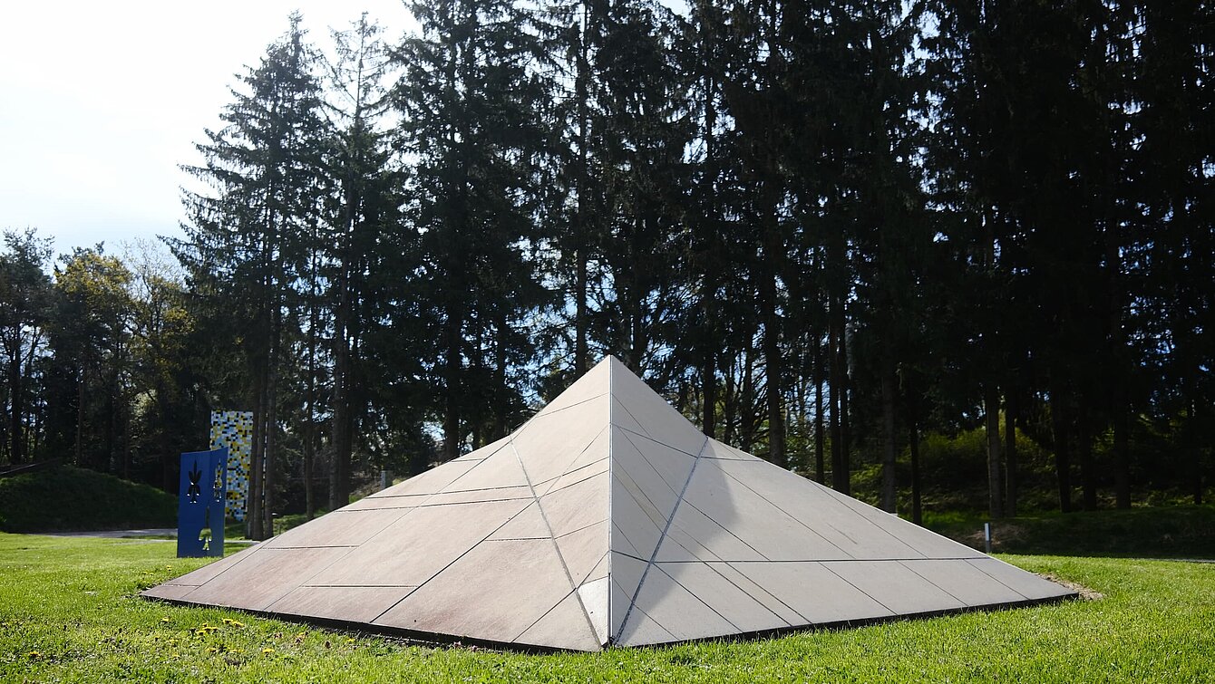 The sculpture in the shape of a stealth ship rests on the meadow. It is covered with beige-colored panels and cannot be detected by radar due to its shape.