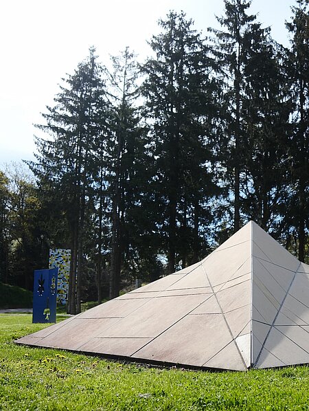 The sculpture in the shape of a stealth ship rests on the meadow. It is covered with beige-colored panels and cannot be detected by radar due to its shape.