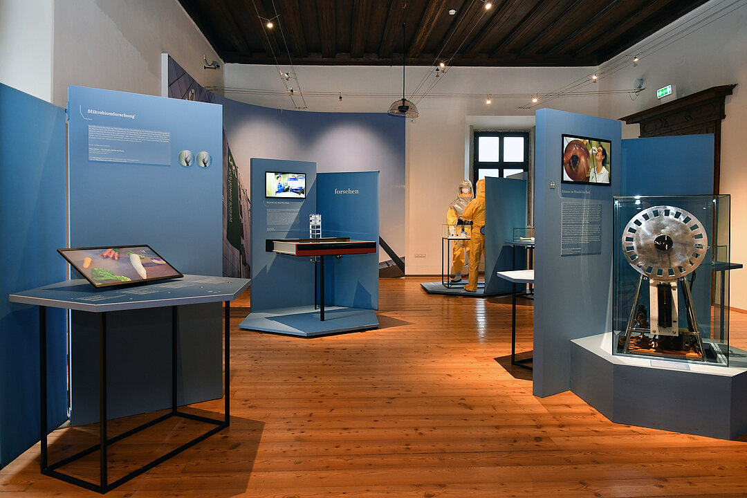 Photo of an exhibition room. In it there are blue text panels and screens on tables.