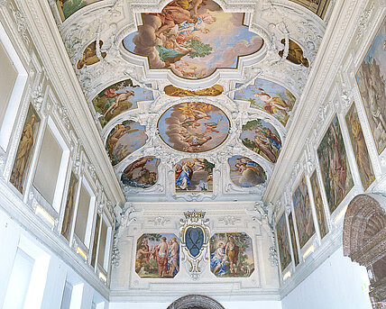 Die Decke einer großen Halle, die mit weißen Stuck und bunten Malereien verziert ist. Die Malereien zeigen Figuren aus Sagen und Religionen. 