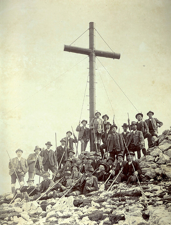 Alte schwarz-weiß Fotografie. Auf einer Bergspitze steht eine Gruppe von Männern in Tracht, um ein großes Gipfelkreuz herum.