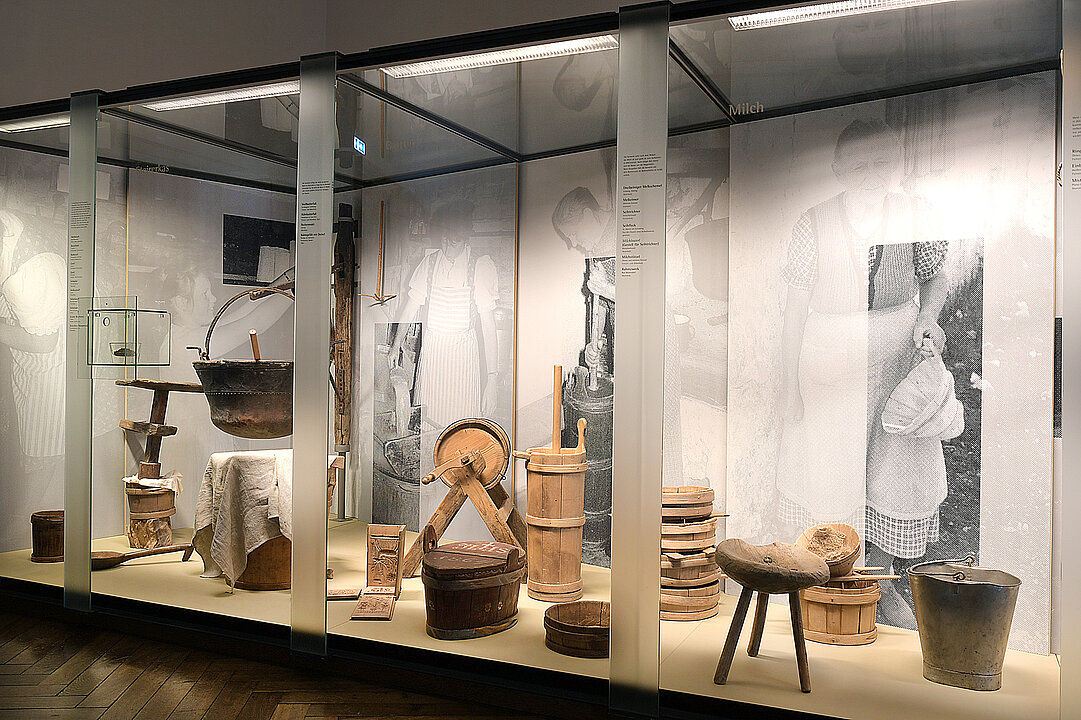 Large glass display case. In it are many objects made of wood, which are needed for milk processing. E.g. buckets, stools and a butter churn.