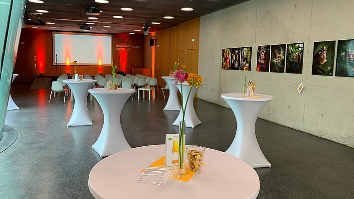 Möbelaufbau für einen Empfang mit Vortrag im Auditorium. Im vorderen Bereich stehen Stehtische mit Dekoration. Im hinteren Bereich ist eine Reihenbestuhlung vor dem Podium. An den Wänden hängen Bilder und das Auditorium ist in stimmungsvolles Licht getaucht.