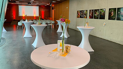 Furniture setup for a reception with lecture in the Auditorium. In the front area are bar tables with decoration. In the back area is a row seating in front of the podium. Pictures hang on the walls and the Auditorium is bathed in atmospheric light.
