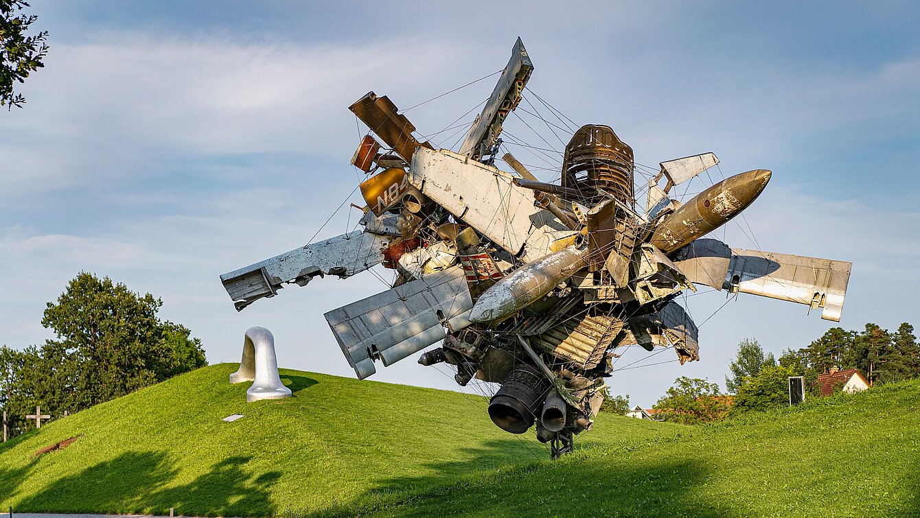 Blick vom Wegesrand auf die Skulptur "Airplane Parts & Hills" von Nancy Rubins und "Die Erdkugel als Koffer" von Peter Weibel in der Ferne