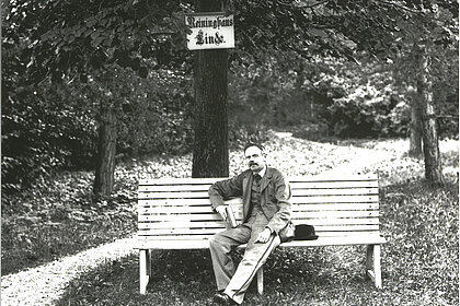 Peter Rosegger sitzt auf einer Bank unter einem Baum, auf dem eine Tafel mit der Aufschrift "Reininghaus Linde" angebracht ist, schwarz-weiß.