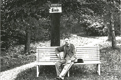 Peter Rosegger is sitting on a bench under a tree, on which a plaque with the inscription "Reininghaus Linde" is attached, black and white.