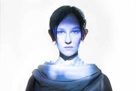 Ein Porträt einer Person mit blauen Augen und kurzen, glatten, dunklen Haaren, die die oder den Betrachtenden ansieht. In der Mitte ist das Porträt stark beleuchtet.