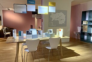 Zu sehen ist der Hauptraum der Ausstellung "Warum?" Im Vordergrund steht ein Tisch mit Informationsmaterial und Faksimile. Im Hintergrund ist eine Landkarte der Steiermark zu sehen.