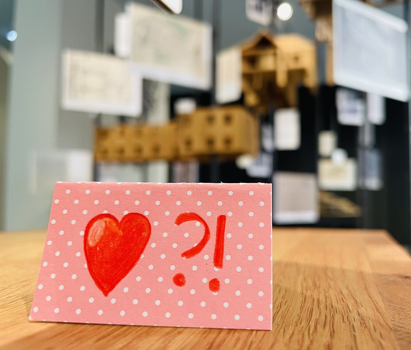 Zu sehen ist ein rosarotes Kärtchen mit einem roten Herz darauf in ersten Ausstellungsbereich. Im Hintergrund sind Museumsmodell aus Holz zu erkennen. 