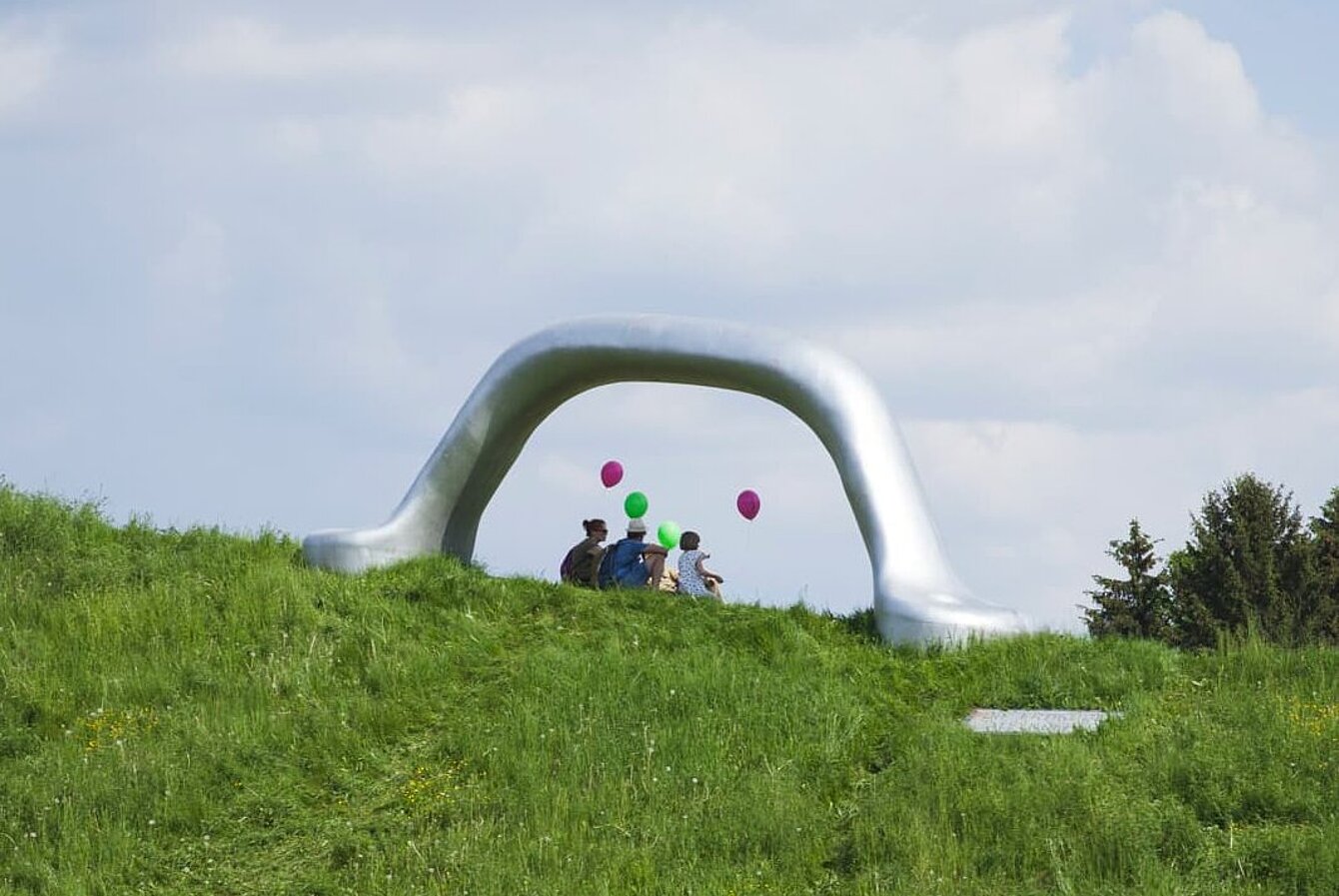 Eine  Gruppe von Besuchern des Frühlingsfest 2023, hat unter dem Kunstwerk  "Die Erdkugel als Koffer" von Peter Weibel Platz genommen. Das Kunstwerk gleicht einem überdimensionalen Koffergriff. 