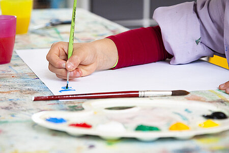 Eine Kinderhand mit Pinsel, die malt. Im Vordergrund ist eine Farbpalette