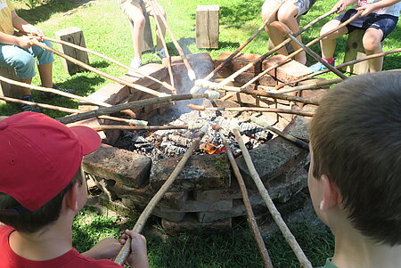 Kinder sitzen auf Holzklötzen rund um eine mit Ziegel begrenzte Feuerstelle und auf  Holzspießen braten sie Würstel und Stockbrot am offenen Feuer