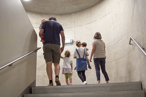 Man sieht mehrere Menschen verschiedenen Alters von hinten, wie sie die Stiegen im Kunsthaus hinaufgehen