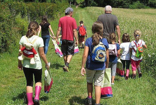 Sammeln, forschen und dokumentieren Kinder und Erwachsene gehen mit Sackerl auf einer Wiese entlang  in Richtung Wald